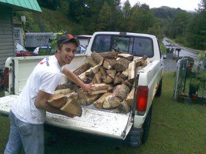 Young man smiling and gesturing towards a pile of firewood loaded in the back of a white pickup truck, parked beside a road.